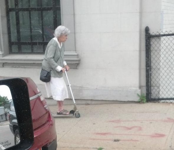 Foto Divertente: Signora con monopattino che corre sul marciapiede